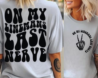 May include: A gray t-shirt with the text "On My Linemans Last Nerve" in black retro-style font on the back. The front of the shirt has a black skeleton hand making a peace sign with the same text in a circle around it.