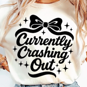 May include: Cream-colored t-shirt with a black bow graphic and the words "Currently Crashing Out" in a playful font. The design includes star accents, creating a casual and fun aesthetic. The shirt is a comfortable, everyday wear.