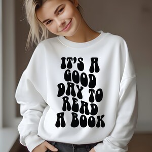 Op de afbeelding: Wit sweatshirt met zwarte tekst die "Het is een goede dag om een boek te lezen" zegt in een retro lettertype.