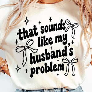 Könnte beinhalten: Cremefarbenes T-Shirt mit dem schwarzen Schriftzug "that sounds like my husband's problem". Das Design umfasst Schleifen-Illustrationen und Stern-Akzente, die eine verspielte und lässige Ästhetik schaffen.