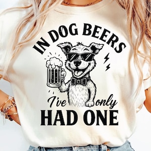 May include: Cream-colored t-shirt featuring a black and white illustration of a dog wearing sunglasses and a bow tie, holding a beer mug. Text reads "IN DOG BEERS I've only HAD ONE".