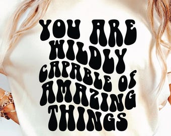 Puede incluir: Camiseta color crema con la frase "YOU ARE WILDLY CAPABLE OF AMAZING THINGS" en letras negras y audaces de estilo retro. La camiseta tiene cuello redondo y mangas cortas. El diseño es casual e inspirador.
