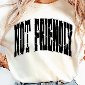 May include: Cream-colored sweatshirt with the text "NOT FRIENDLY" in bold, distressed black letters. The text is centered on the front of the shirt. The shirt has a crew neck and long sleeves.