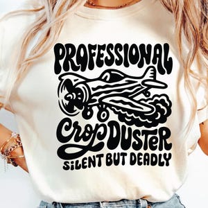May include: Cream-colored t-shirt featuring a black graphic of a crop duster plane with the text "PROFESSIONAL CROP DUSTER SILENT BUT DEADLY" in a retro font. The design is centered on the shirt.