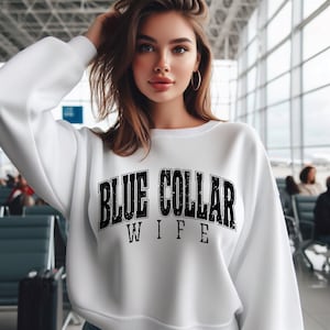 May include: A white sweatshirt with the text "BLUE COLLAR WIFE" in a black, distressed font. The sweatshirt has a crew neck and long sleeves. The image is taken indoors, with a blurred background.