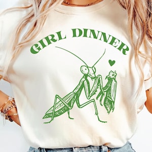 May include: Cream-colored t-shirt with a green graphic of two praying mantises and the words "GIRL DINNER" above them. A small green heart is above the mantises. The graphic is a whimsical illustration.