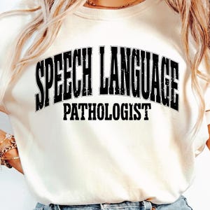 Puede incluir: Sudadera color crema con la frase "SPEECH LANGUAGE PATHOLOGIST" impresa en letras negras gruesas y desgastadas. El texto está arqueado en la parte delantera. Una prenda informal para el uso diario.