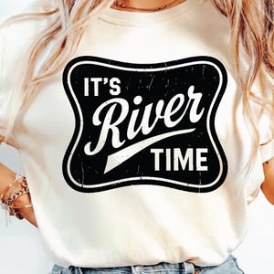 Può includere: Una maglietta color crema con una grafica in bianco e nero. La grafica è di forma rettangolare con angoli arrotondati e il testo "IT'S River TIME" è stampato in grassetto. La maglietta è di stile casual.