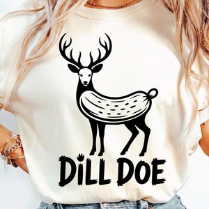 Puede incluir: Camiseta color crema con un gráfico negro de un ciervo con cuerpo en forma de pepinillo. El texto "DILL DOE" está impreso en negrita, en negro, debajo del ciervo. El diseño es una versión lúdica del tema del ciervo y el pepinillo.