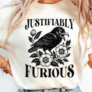 May include: Cream-colored t-shirt featuring a black illustration of a crow with an open beak, surrounded by flowers and leaves. The words "JUSTIFIABLY" and "FURIOUS" are printed above and below the crow design in a bold, black font.