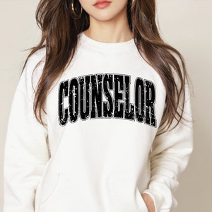 Op de afbeelding: Wit sweatshirt met het woord "COUNSELOR" gedrukt in zwarte, verweerde letters.