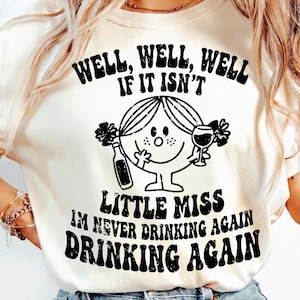 May include: Cream-colored t-shirt with a black graphic of a Little Miss character holding a wine glass and a bottle. The text on the shirt reads "WELL, WELL, WELL IF IT ISN'T LITTLE MISS I'M NEVER DRINKING AGAIN DRINKING AGAIN."