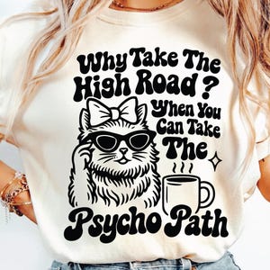 Puede incluir: Camiseta color crema con un gráfico negro de un gato con gafas de sol y un lazo, levantando un dedo y una taza de café. El texto dice: "Why Take The High Road? When You Can Take The Psycho Path."