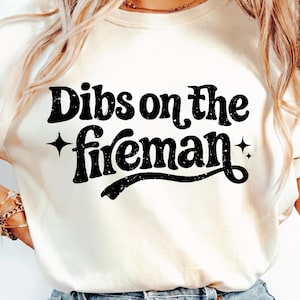 Puede incluir: Camiseta color crema con la frase "Dibs on the fireman" en letras negras desgastadas. El texto está acentuado con gráficos de estrellas, creando una estética vintage.