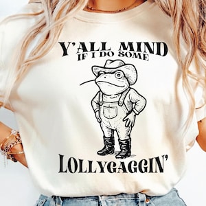 Puede incluir: Camiseta color crema con un dibujo de una rana de dibujos animados con sombrero de vaquero y peto. El texto "Y'all Mind If I Do Some Lollygaggin'" está escrito. La rana es en blanco y negro, con un objeto fino en la boca.