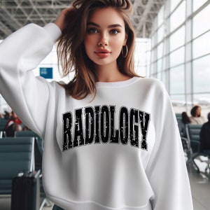 Może przedstawiać: Biała bluza z napisem "RADIOLOGY" wydrukowanym dużymi, postarzanymi czarnymi literami na piersi. Bluza ma okrągły dekolt i długie rękawy. Tło to rozmyty terminal lotniskowy.