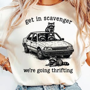 May include: Cream-colored t-shirt featuring a black and white illustration of a car with raccoons. The text reads "get in scavenger" above the car and "we're going thrifting" below. The graphic depicts raccoons in and on top of the car.