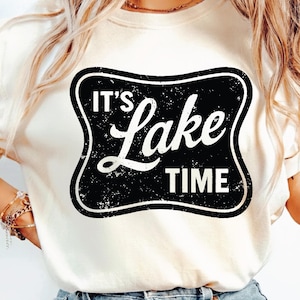 Puede incluir: Camiseta color crema con un gráfico en blanco y negro que dice "IT'S Lake TIME". El gráfico tiene un estilo desgastado, con un borde negro y letras blancas. La camiseta es de estilo casual.