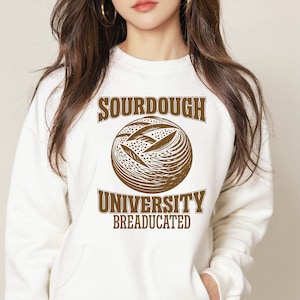 Può includere: Felpa bianca con testo marrone che recita "Sourdough University Breaducated" e un'illustrazione marrone di una pagnotta di pane a lievitazione naturale.