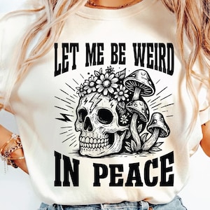 以下が含まれることがあります： クリーム色のTシャツ。黒いグラフィックで、花の冠とキノコをつけた骸骨が描かれています。「LET ME BE WEIRD IN PEACE」の文字が骸骨のデザインの上と下にプリントされています。カジュアルウェアです。