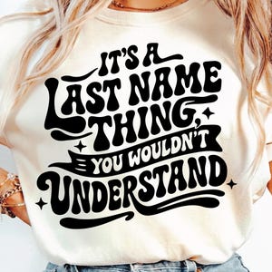 May include: A cream-coloured t-shirt featuring the text "IT'S A LAST NAME THING, YOU WOULDN'T UNDERSTAND" in bold, black, retro lettering. The design incorporates stars and decorative elements, giving a vintage feel.