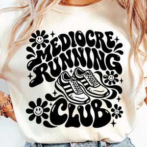May include: A cream-colored t-shirt featuring a black graphic print with the text "MEDIOCRE RUNNING CLUB" in a bold, wavy font. The design includes two running shoes in the center, surrounded by four stylized flowers with smiley faces.