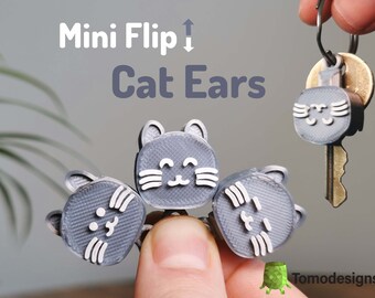May include: Three grey cat ear keychains with white smiling faces. The keychains are attached to a keyring and a key. The text "Mini Flip Cat Ears" is visible in the image.