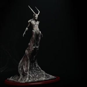 May include: A tall, slender, grey-toned humanoid figurine with two long, curved horns extending from its head. The figure features a textured, organic, ribbed body that flares out at the base, standing on a circular, dark red, elevated platform.