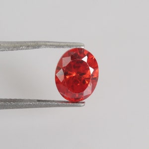 May include: A round, faceted, red gemstone held by tweezers. The gemstone is a vibrant orange-red color and has a high degree of brilliance.