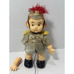 May include: Vintage doll in a military-style uniform. The doll has a gray velvet jacket with gold trim, black boots, and a matching hat with red feathers. One arm is detached. The doll's face is painted with rosy cheeks and a sweet expression.