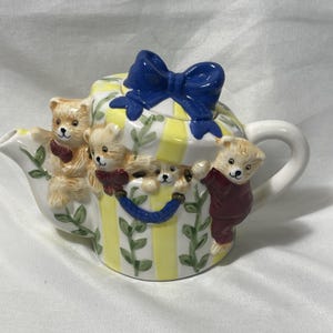 May include: A whimsical ceramic teapot featuring a yellow and white striped design with a blue bow on the lid. The teapot is adorned with several teddy bear figurines, adding a playful touch to the design. The teapot has a white handle and spout.