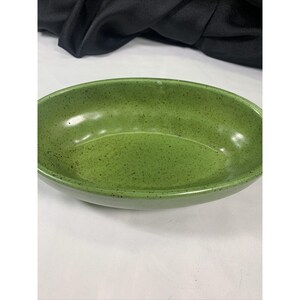 May include: A speckled green ceramic oval serving bowl. The bowl has a glossy finish and a shallow, elongated shape. The interior and exterior are the same shade of green with dark speckles. The bowl is empty and sits on a white surface.