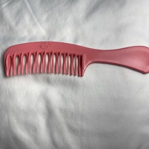 May include: A pink plastic comb with a wide handle and widely spaced teeth. The comb has a curved design and a smooth finish. The brand name is visible on the top of the comb.