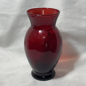 May include: A ruby red glass vase with a flared top and a rounded body. The vase has a dark base and a smooth, glossy finish. The vase is approximately 8 inches tall.