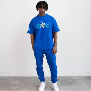以下が含まれることがあります： 青いTシャツを着た男性。Tシャツには、青と緑のロゴで「Sega」、青と白のロゴで「Sonic」と書いてある。彼は、ロイヤルブルーのスウェットパンツと白いスニーカーも履いている。