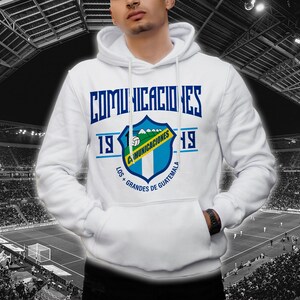 Puede incluir: Sudadera con capucha blanca con un escudo azul y verde con el texto "Comunicaciones" y "Los + Grandes de Guatemala". El escudo también incluye un balón de fútbol y montañas. El texto "1919" aparece a ambos lados del escudo.