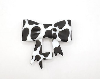 May include: A black and white bow with a cow print design. The bow is made of a soft material and has a classic bow shape. The bow is a fun accessory for any outfit.