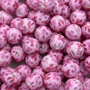 May include: A collection of pink round beads with a bow pattern in varying shades of pink. The beads are clustered together, showcasing their decorative design. These beads are ideal for jewelry making and crafting projects.
