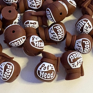 May include: A collection of brown, football-shaped beads with the text "Pot Head" and "Head" in white lettering. The beads have a hole for stringing and are scattered across a light surface.