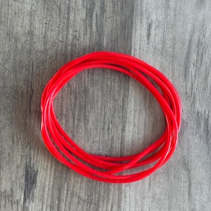 May include: Bright red, flexible tubing coiled in a circular shape. The tubing is smooth and appears to be made of a pliable material. The background is a gray, wood-grain surface.