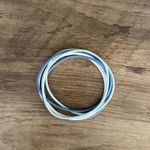 May include: A set of three thin, circular bangles in varying shades of blue and white. The bracelets are smooth, flexible, and feature a matte finish, with one bangle in a light sky blue, one in a medium cornflower blue, and one in a crisp white.