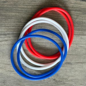 May include: Three circular bangle bracelets in solid colors of red, white, and blue. Each bracelet features a smooth, rounded surface and a simple, continuous loop design. The bracelets are stacked slightly overlapping each other.