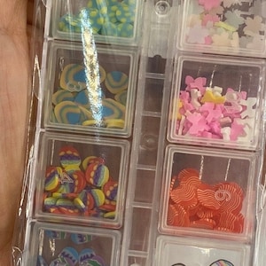 May include: A clear plastic storage box with 10 compartments filled with colorful sprinkles and decorative shapes. The sprinkles are in various colors, including red, white, blue, yellow, green, and orange. The shapes include hearts, stars, circles, and other designs.