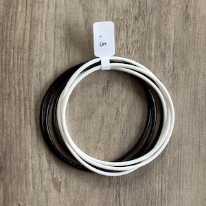 May include: A set of three round bracelets: one black and two white. The bracelets are held together with a white tag that reads "8". The bracelets are on a wooden surface.