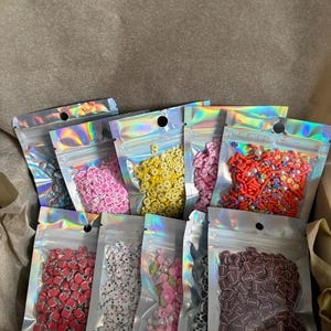 May include: A collection of nine small, clear, holographic resealable pouches filled with various colorful polymer clay slices and beads. The contents include shapes like pink flowers, yellow daisies, red footballs, white playing cards, and small pearls.