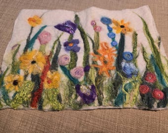 以下が含まれることがあります： 白いフェルトの壁掛けで、刺繍された花の模様が施されています。花は黄色、オレンジ、紫、青、ピンクの色合いです。背景は白です。