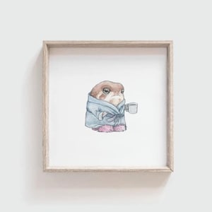 May include: A square wooden frame containing a watercolor illustration of a brown toad wearing a light blue bathrobe and pink slippers, holding a small white mug. The toad has a sleepy expression and is centered on a plain white background.