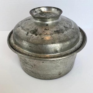 May include: A vintage silver metal pot with a lid. The pot has a worn, aged look and is made of a single piece of metal.