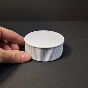 Op de afbeelding: Een witte, ronde container met een geribbelde, cilindrische body en een platte deksel. De container wordt door een hand vastgehouden, wat het ontwerp benadrukt. Het object staat tegen een donkere achtergrond.