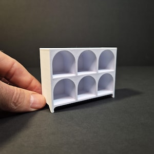 May include: A small white rectangular shelf unit featuring six individual cubby compartments arranged in two rows of three. Each compartment has a rounded arched top design. The structure is made of a solid material with a visible 3D printed texture.